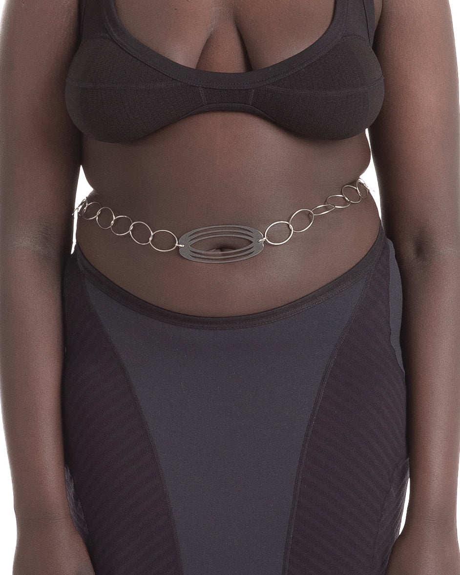 A silver-colored unisex belt with a laser-cut pendant, logo tag, and oval hook, worn around the waist.